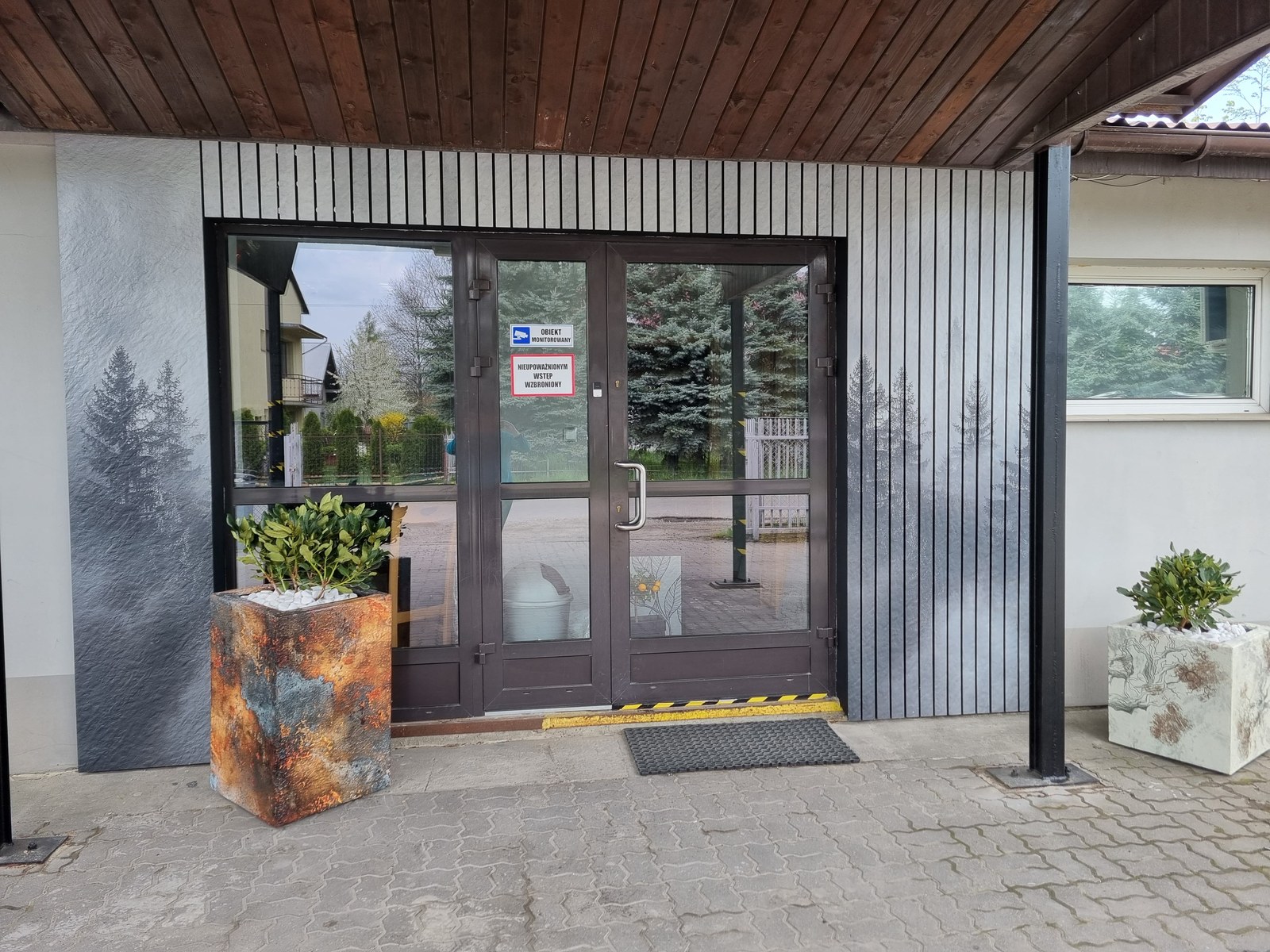 Porównanie grafiki SmartFresk DTS na płycie elewacyjnej i lamelach — motyw leśny na wejściu do budynku, dwie donice kompozytowe